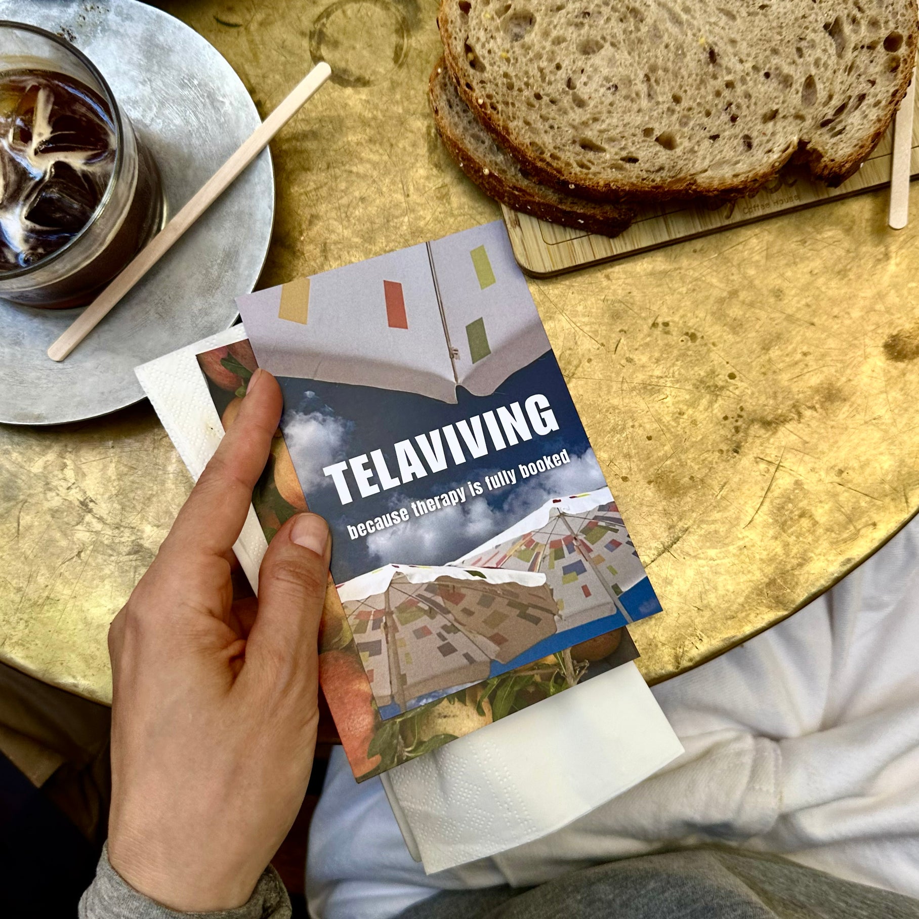 Person holding a Tel Aviv Postcard on a table with coffee and bread, showcasing Tel Aviv's vibrant culture.