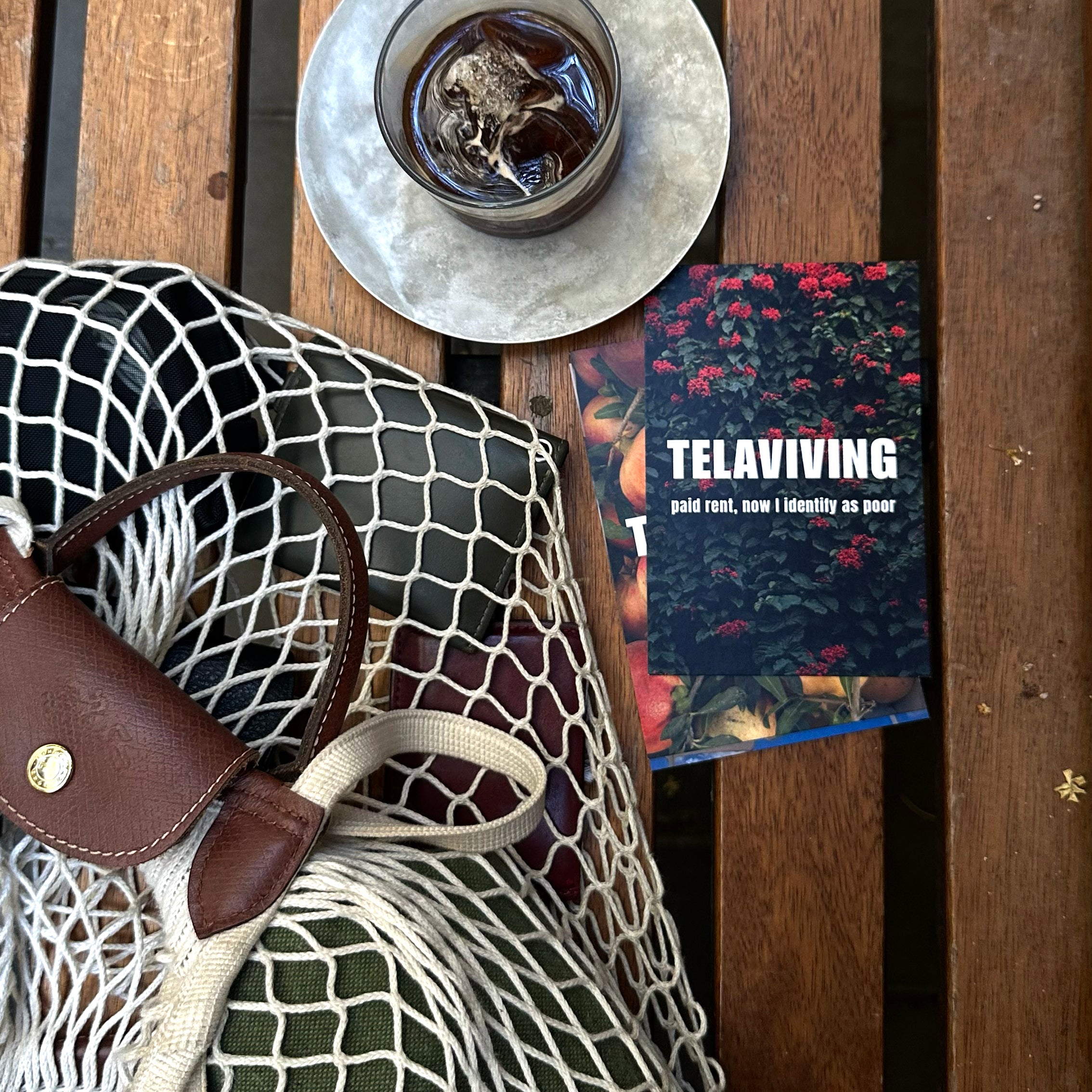 Tel Aviv Postcard featuring vibrant flowers and an artistic title on a wooden table with a drink and bag.